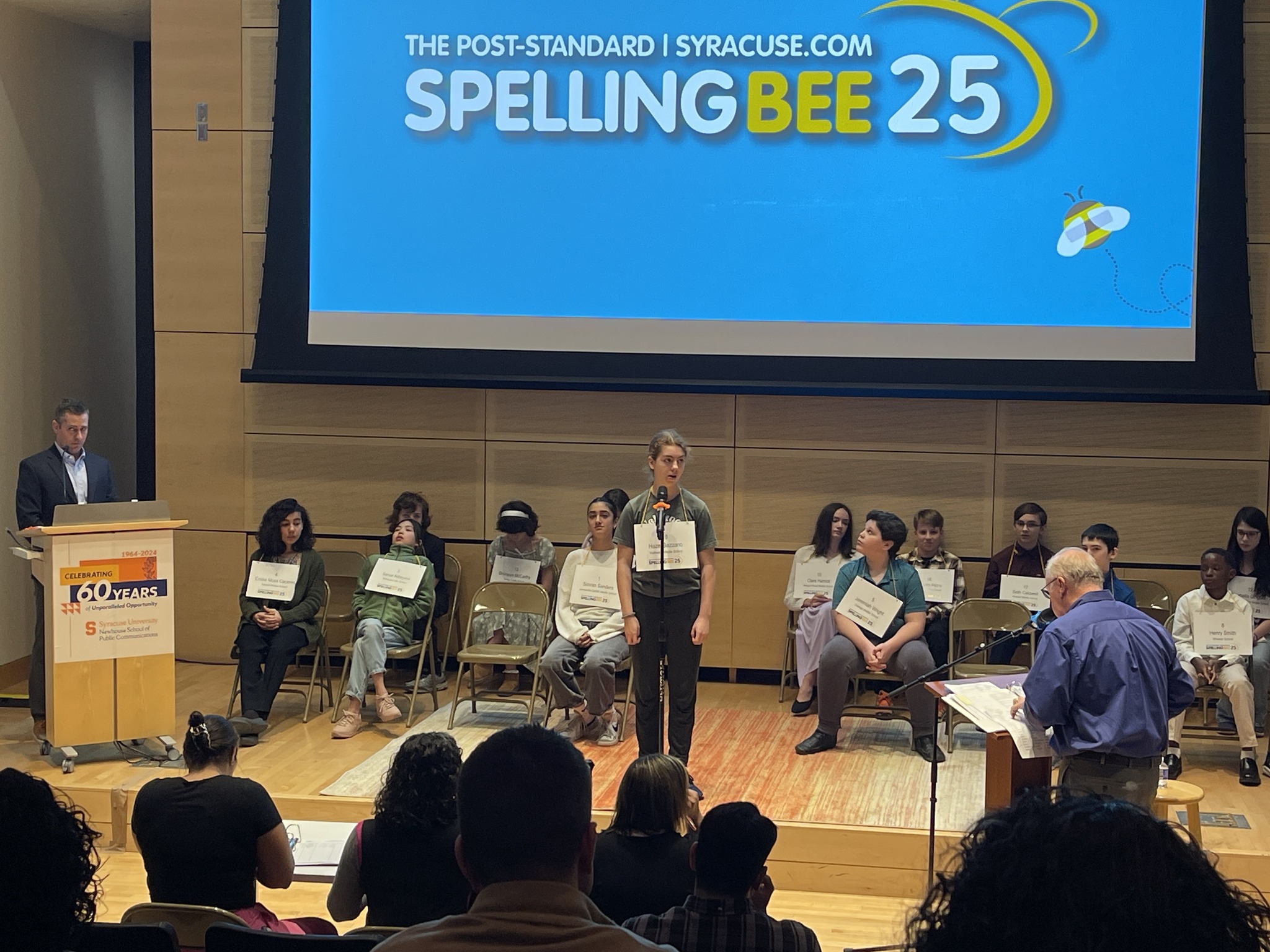 A middle school student stands on a stage with a microphone as others look on.