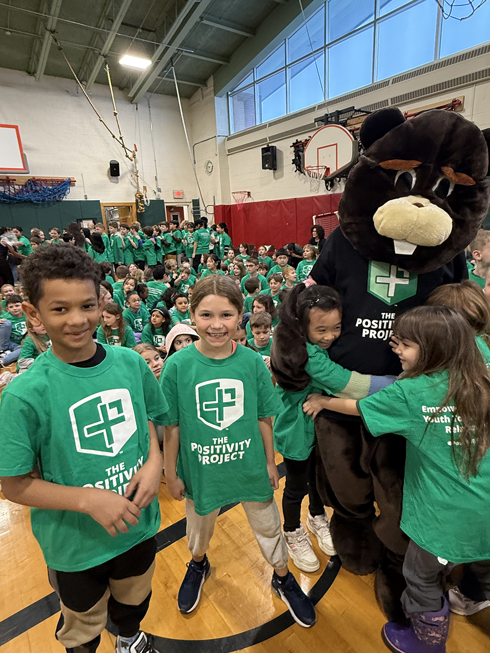 Two elementary aged children hug a giant bear mascot while two others wearing tshirts that say "Positivity Project" look on.