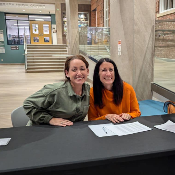 Two HSA members smile warmly seated at a table in a school foyer.