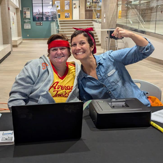 Two HSA parents wearing read headbands sit behind a laptop and a cashbox in the school foyer.