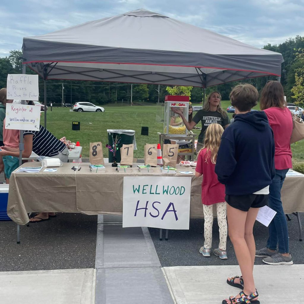 Three people gather in front of a tent labeled as "Wellwood HSA" to listen to an HSA volunteer speaking.