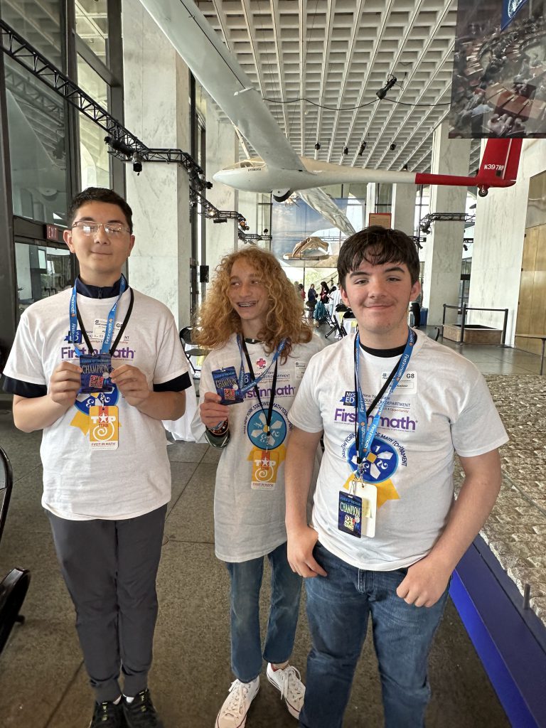 Three middle school students pose with badges around their necks in a convention center setting.