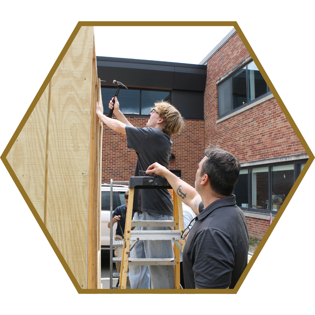 student standing on a ladder hammering a structure while a teacher stands nearby