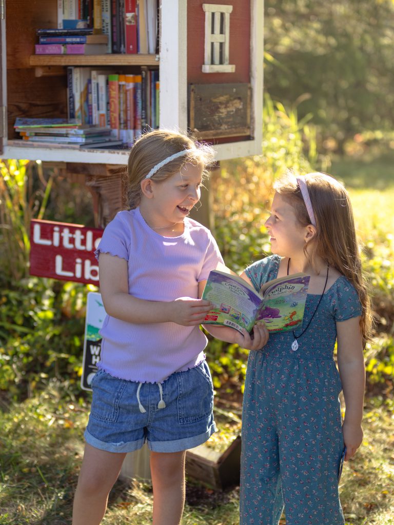 Two girls read book next to little library.