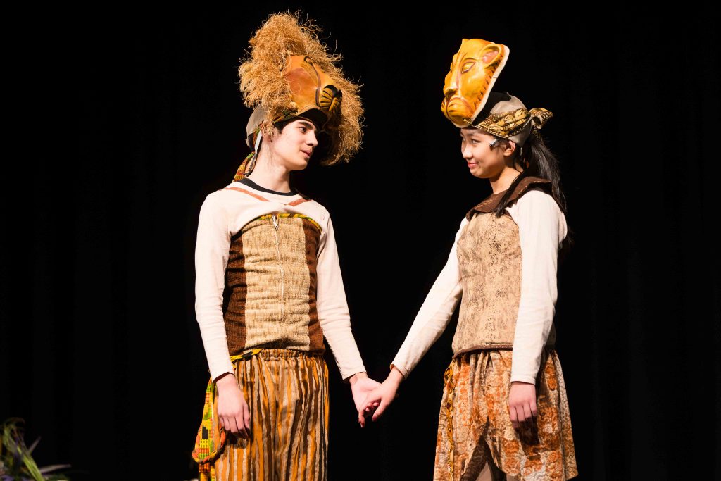 Two students hold hands and look at each other wearing lion costumes in stage performance. 