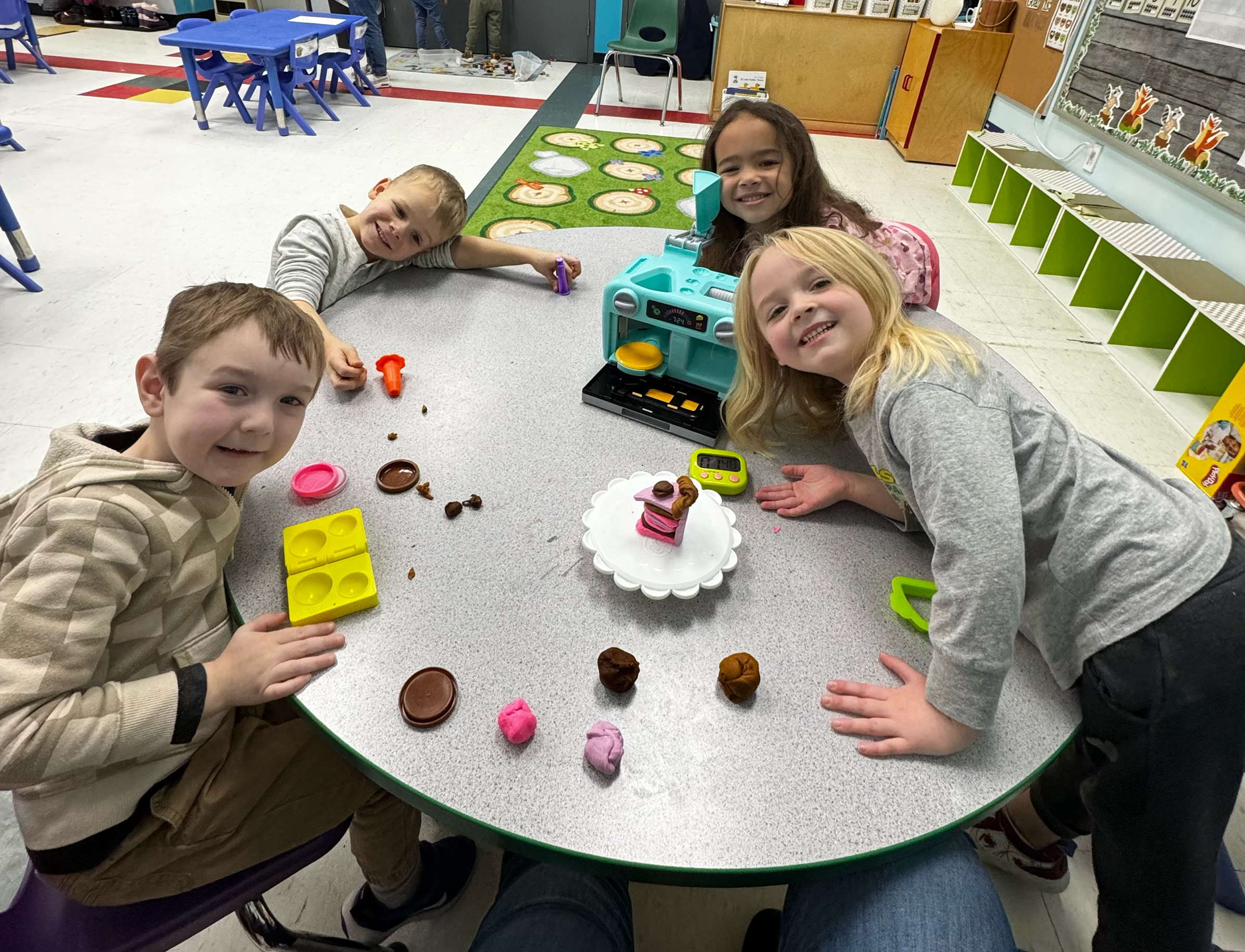 Four preK students seated around a round table, playing with Play-Do. They are leaning in on the table and smiling at the camera.
