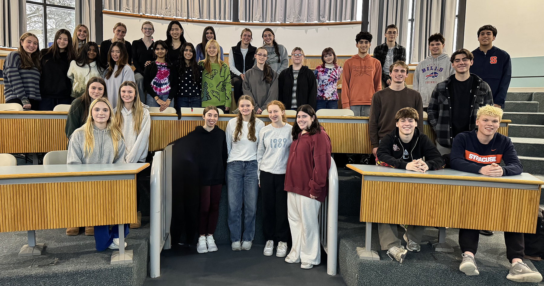 About 40 students seated and standing in a group in a classroom all facing the camera and smiling.