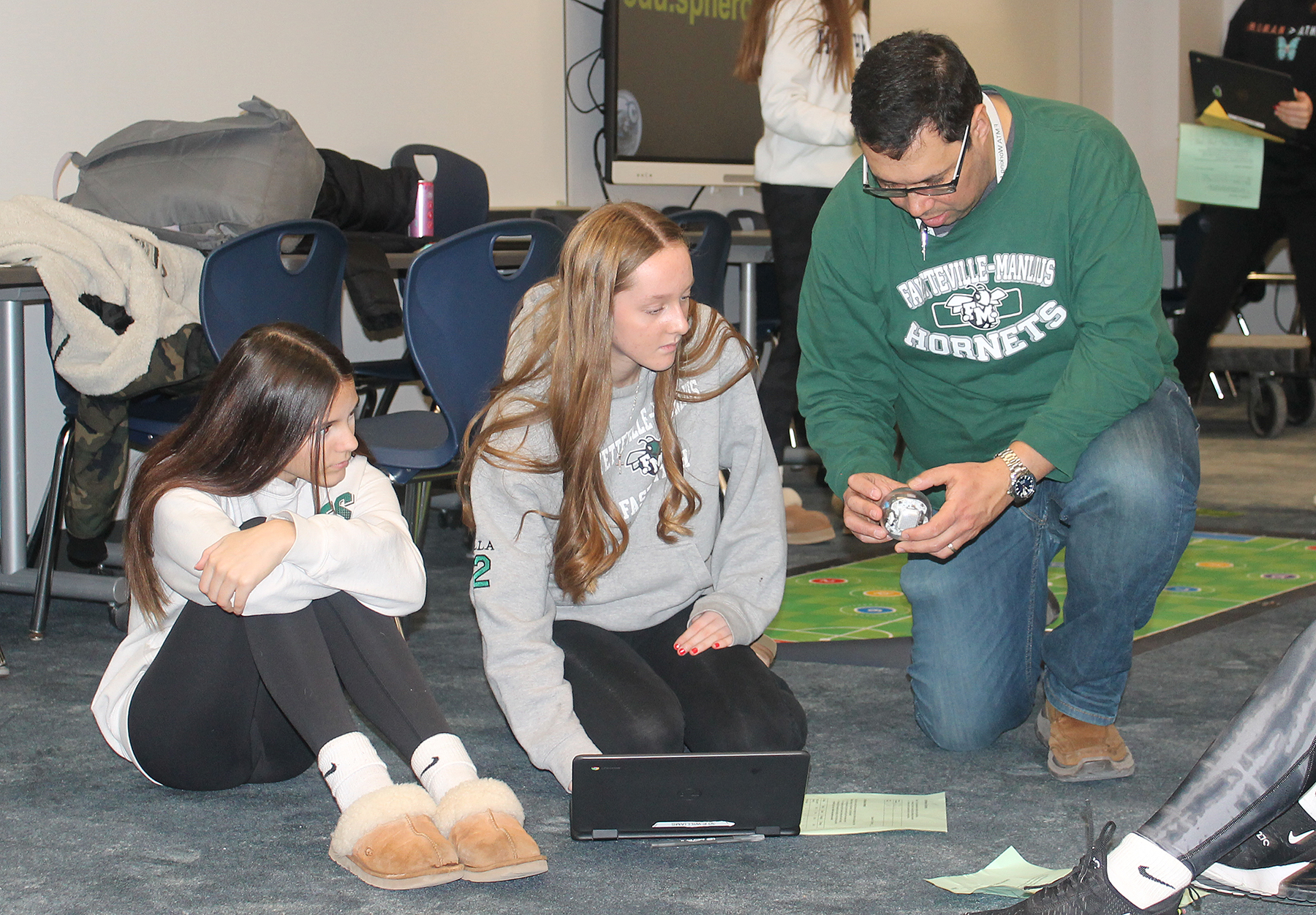 Teacher kneeling to the left of two students, both looking at a sphere the teacher is holding
