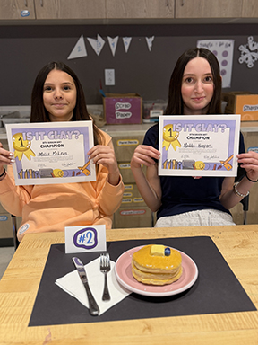 Two girls seated holding certificates with heir winning pancake sculpture on the table in front of them