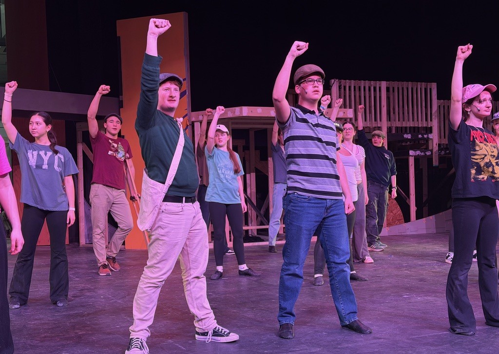 Students stand on stage with one arm raised while practicing Newsies musical.