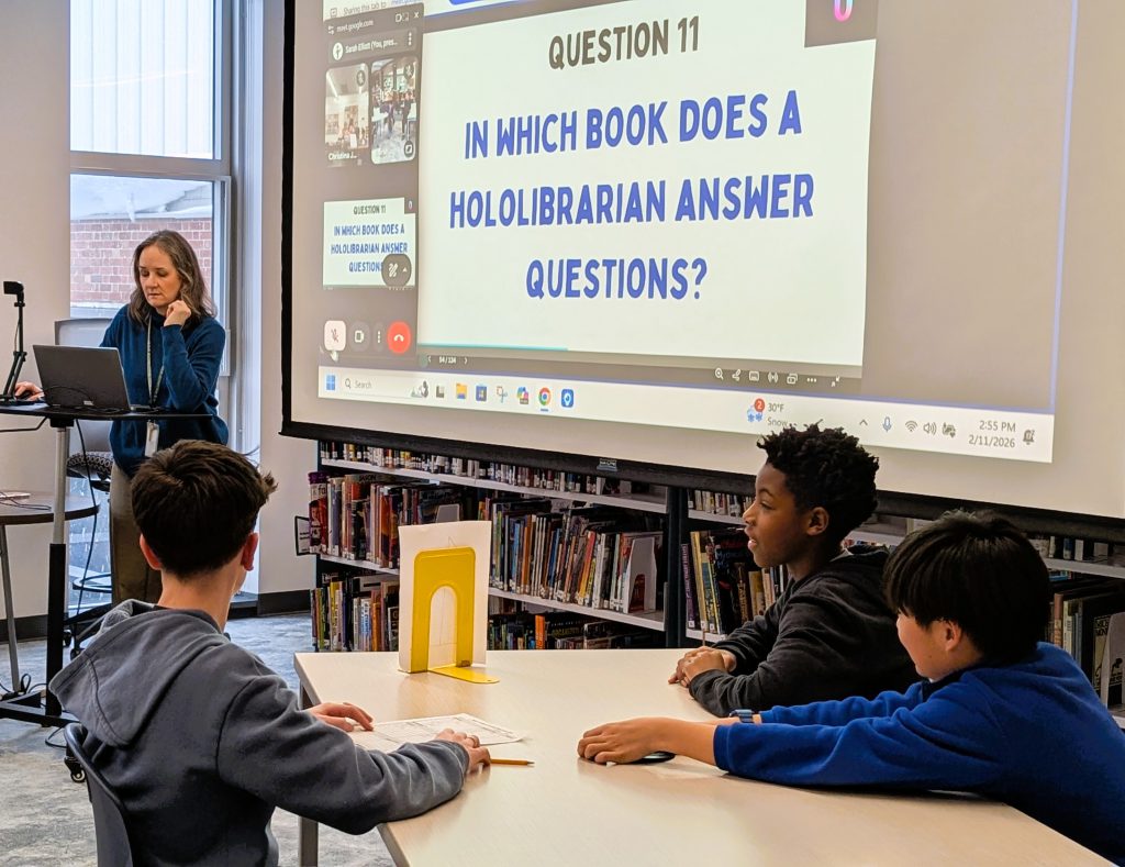 Librarian Sarah Elliott stands at podium while students sit at table during Battle of the Books competition.