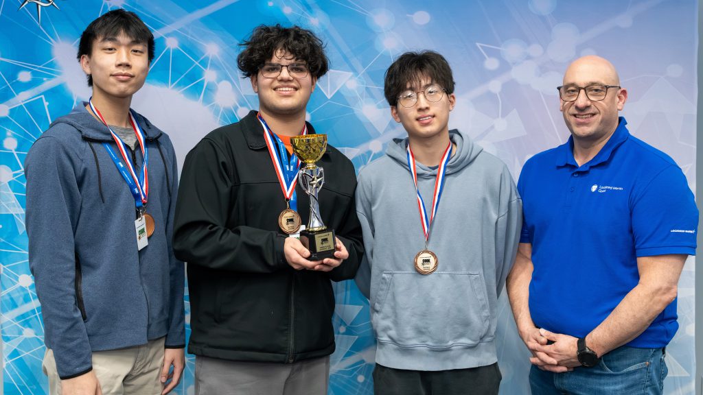 Three students and a teacher standing side by side. The students are holding medals and a trophy.
