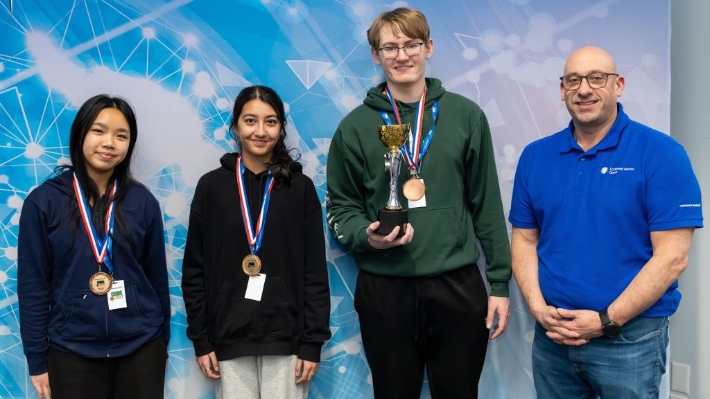 Three students and a teacher standing side by side; the students are wearing medals around their necks and one student is holding a trophy