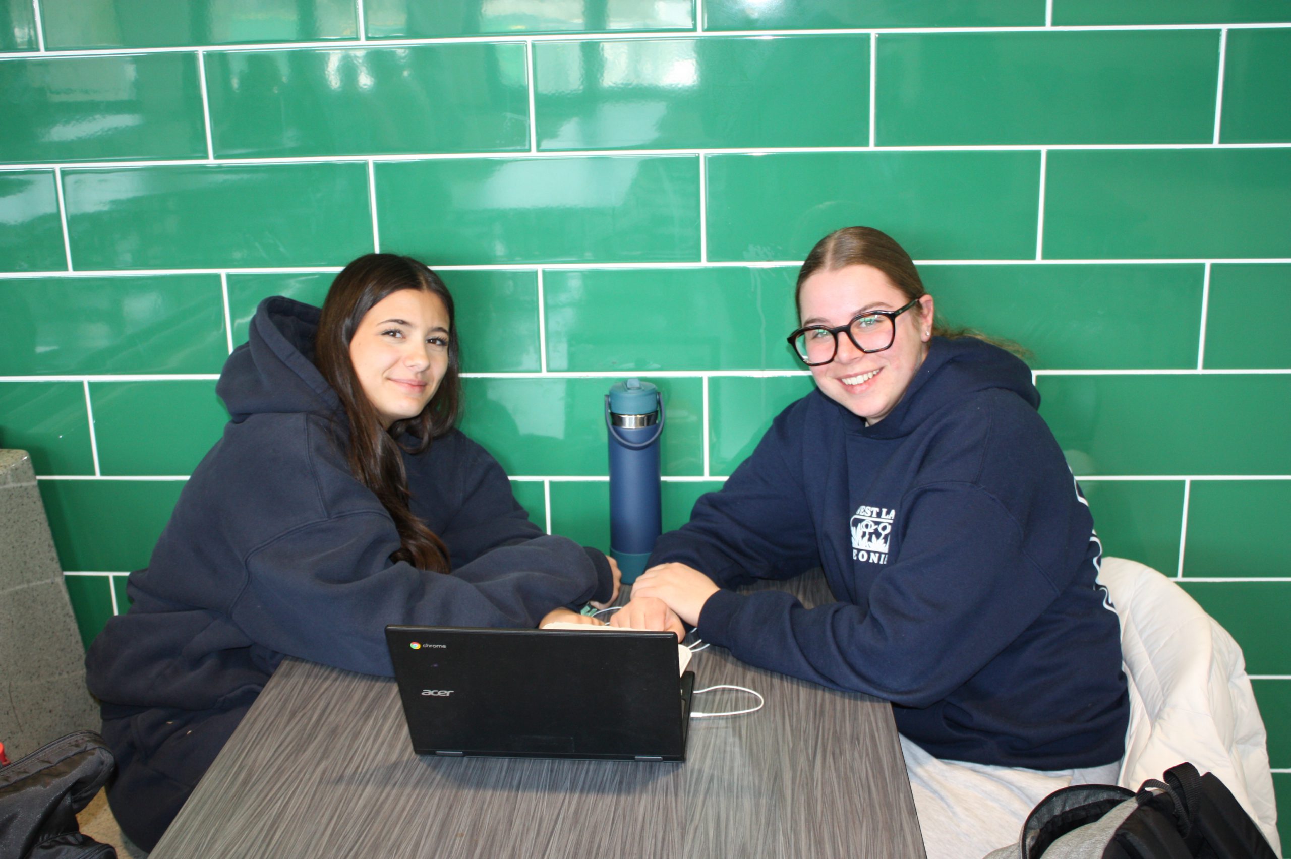 Two girls seated is a botth, leaning towards each other with a laptop on the table between them.
