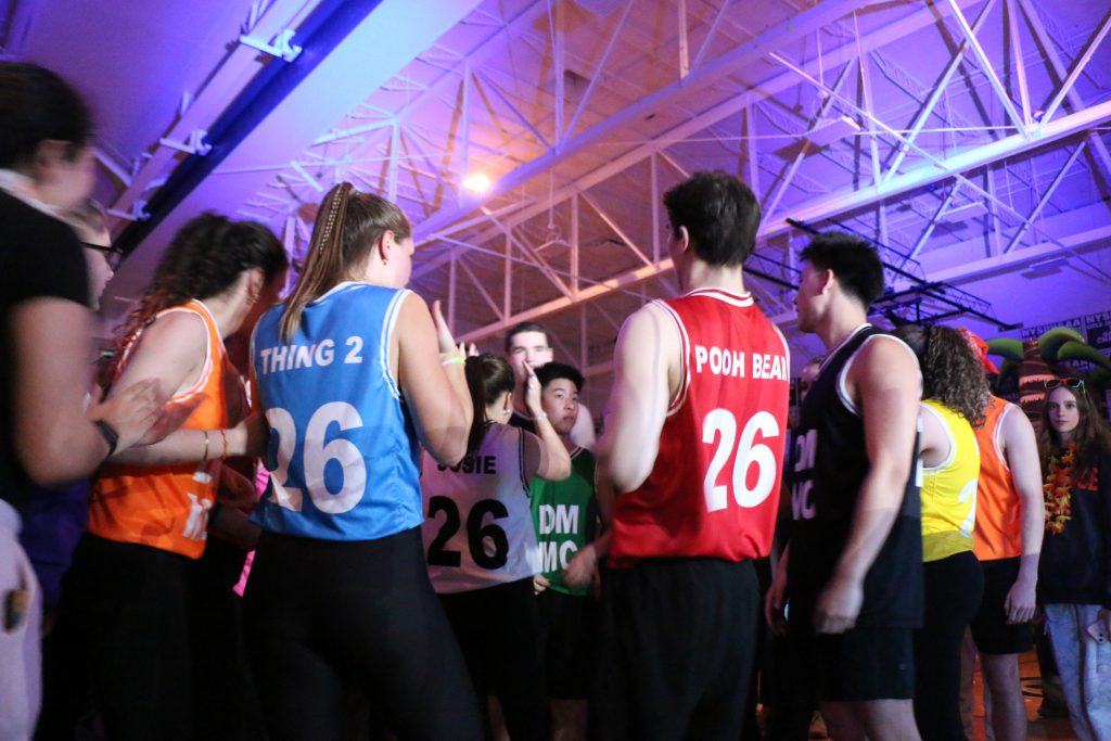 Students dance in darkened gym with lights, wearing colorful sports tank tops. 