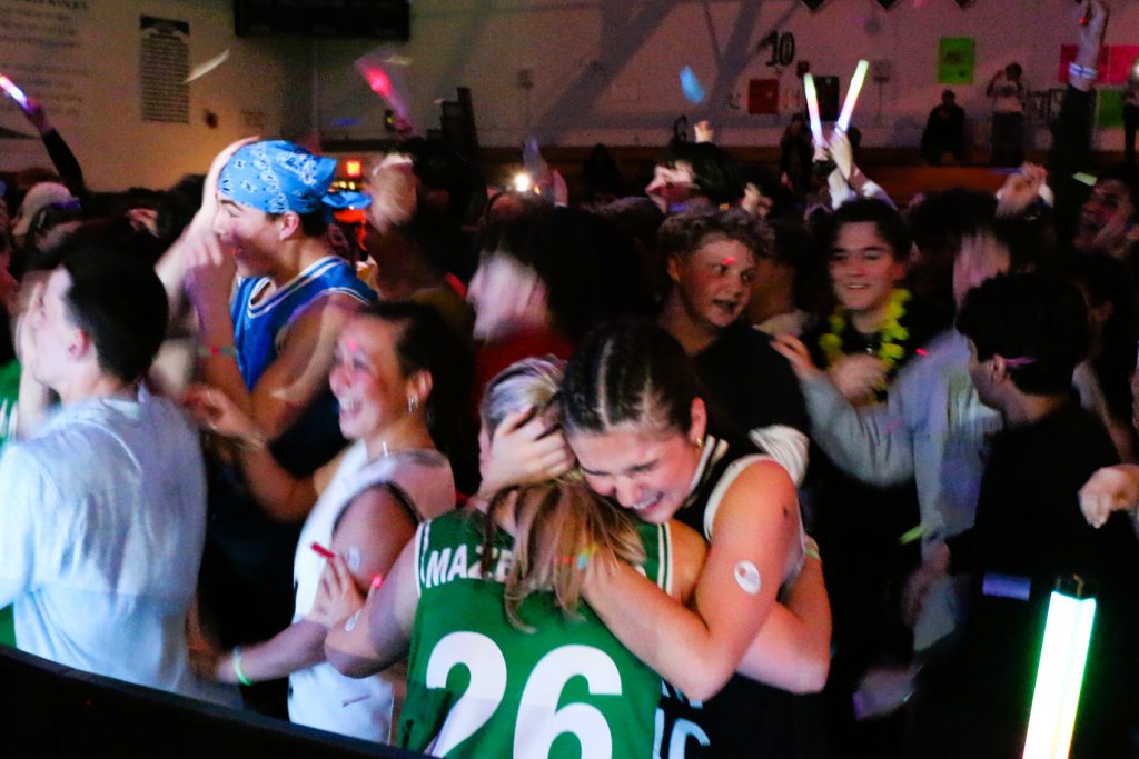 Two students hug in the foreground, a large group of students dance in the background.