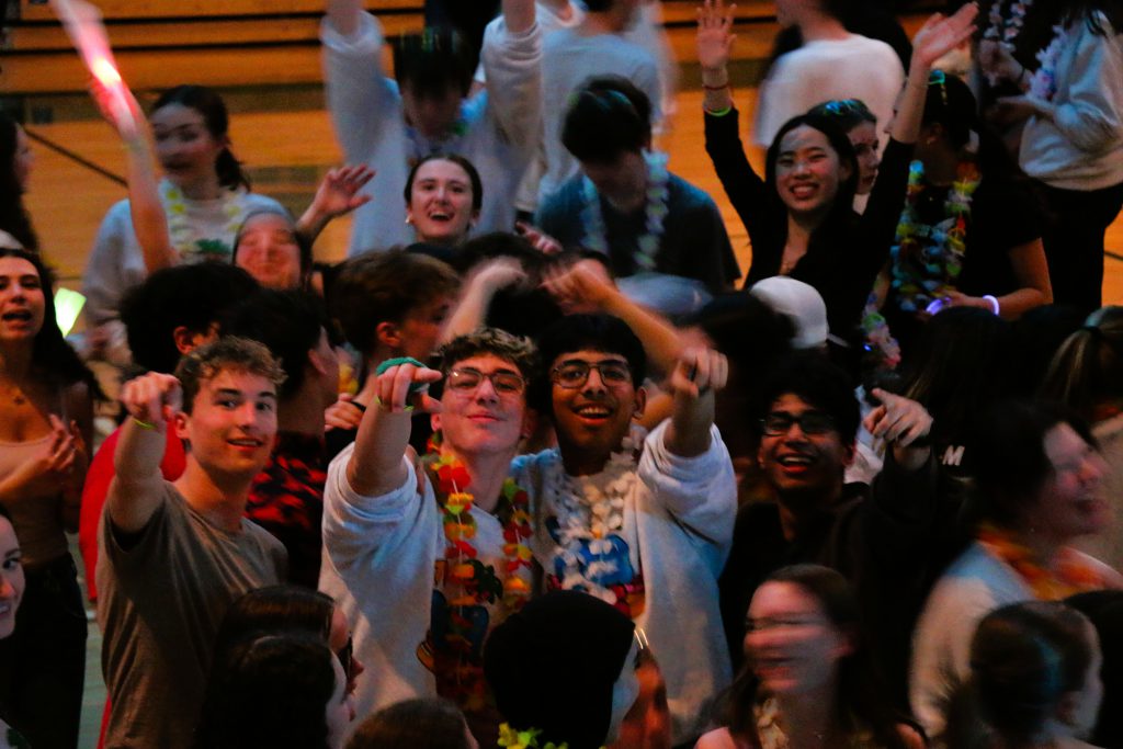 Students smile and point at camera in packed group dancing at Dance Marathon in school gym.