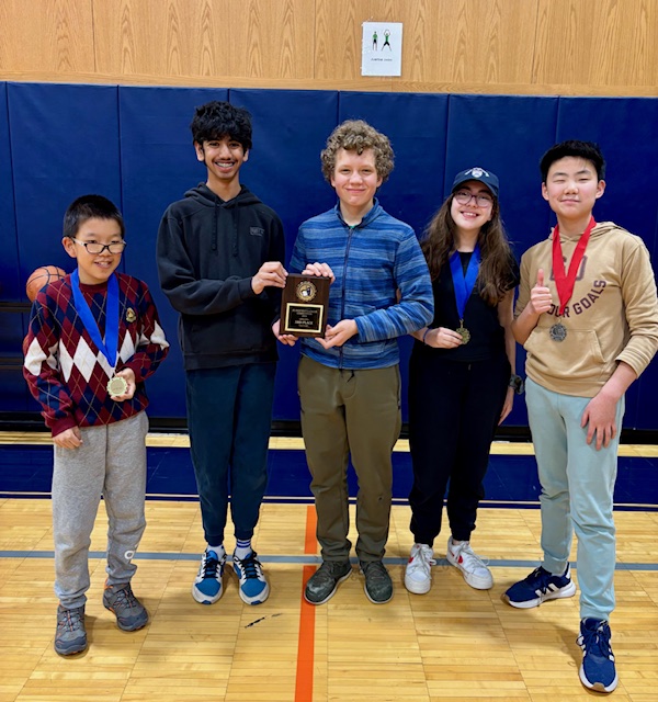 Students stand in line posing for group photo with plaque in front of blue gym mats.