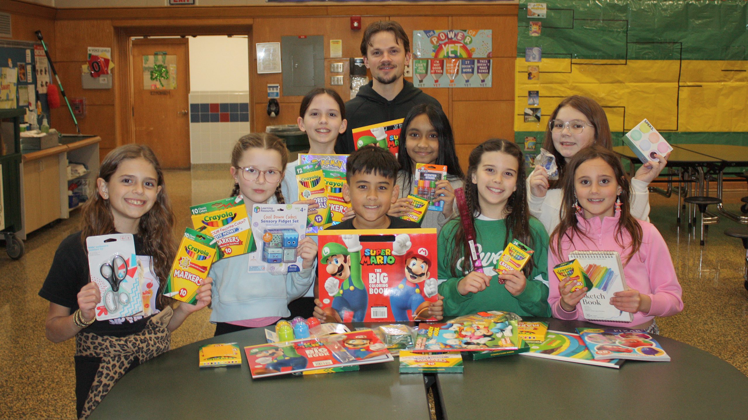 group of students standing behind a table piled with art supplies. Students holding supplies as well
