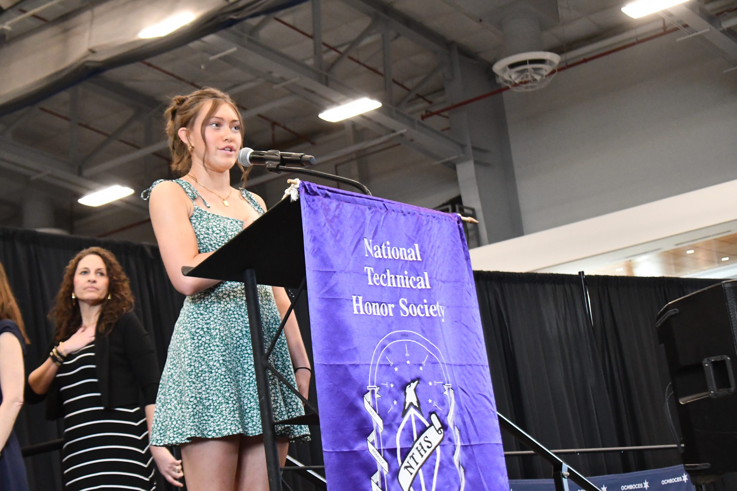Student wearing green dress speaks at podium with National Technical Honor Society banner.
