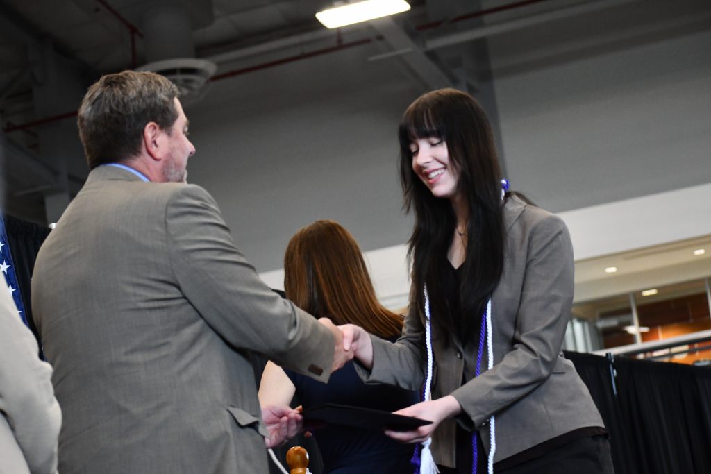 Student, wearing purple and white cords, shakes hands with administrator.
