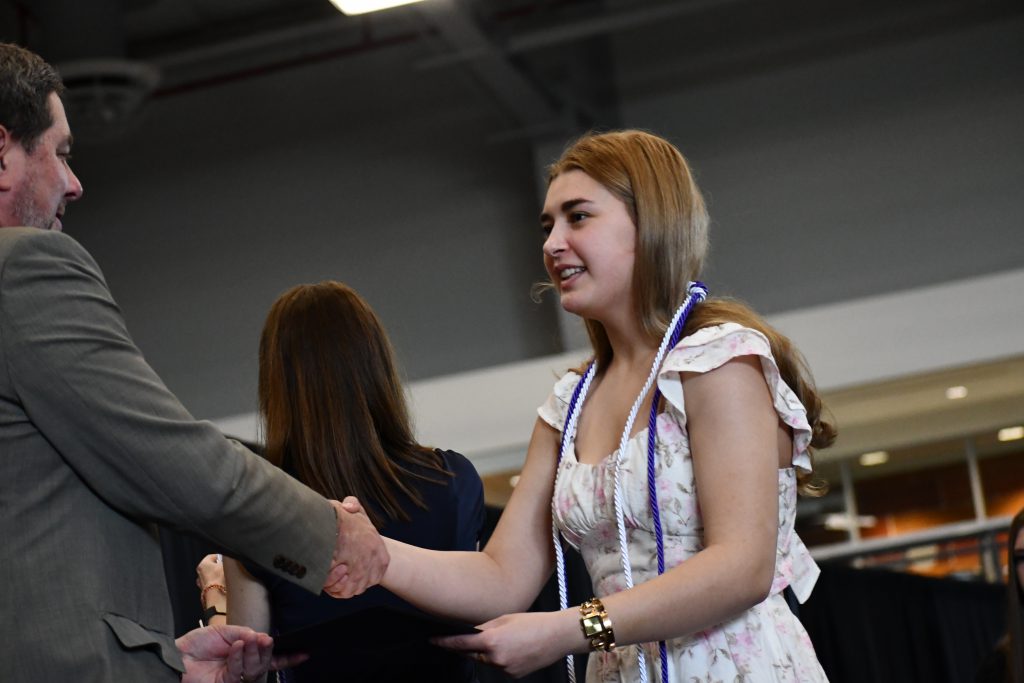 Student, wearing purple and white cords, shakes hands with administrator.