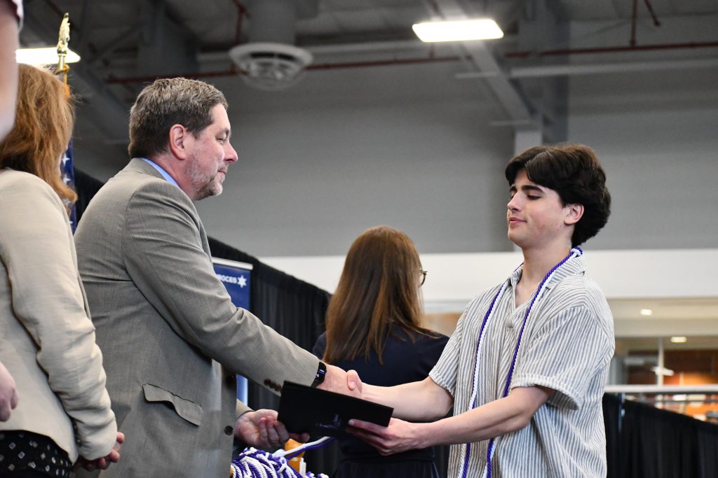 Student, wearing purple and white cords, shakes hands with administrator.