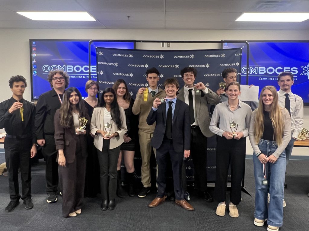 Group of students pose for photo holding medals and ribbons in front of OCM BOCES monitors and logo background.