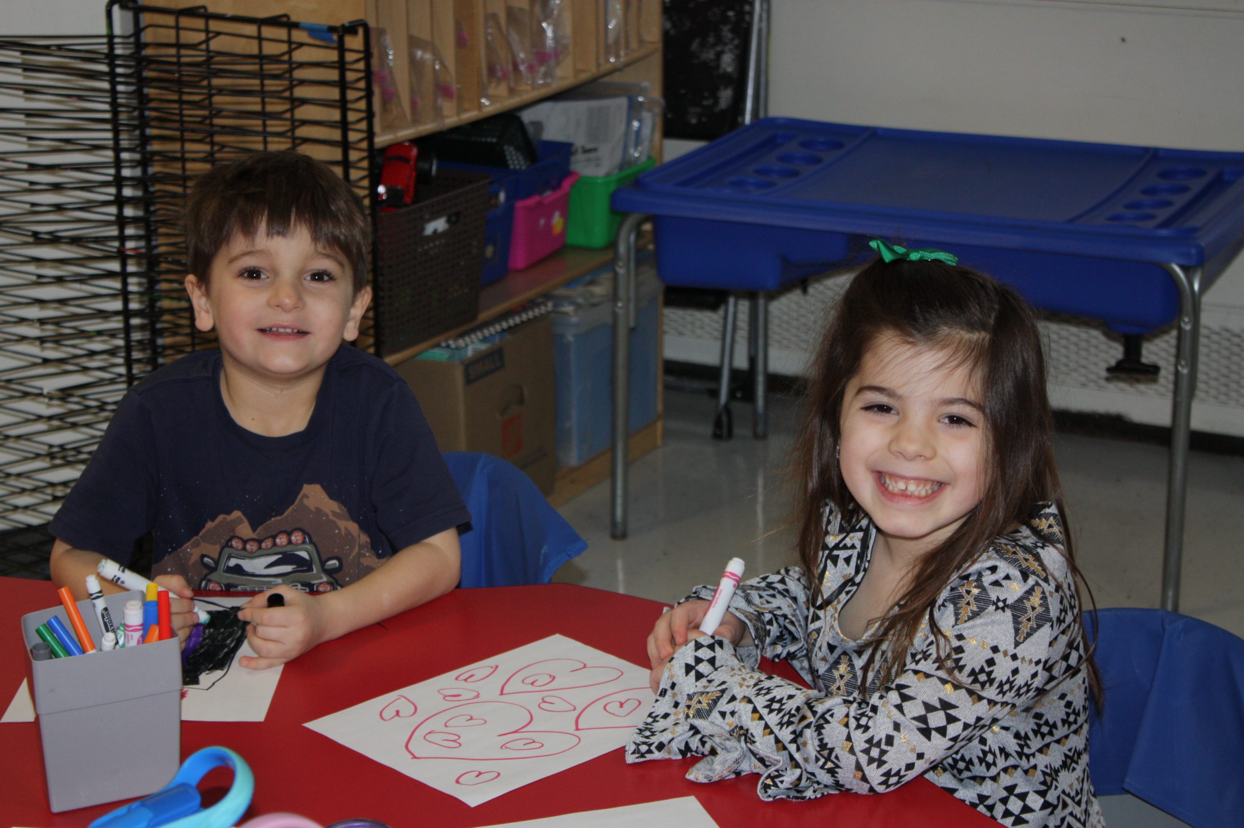 Two students seated at a table drawing and smiling at the camera