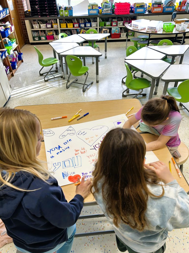 Three students work on poster while sitting at classroom table.