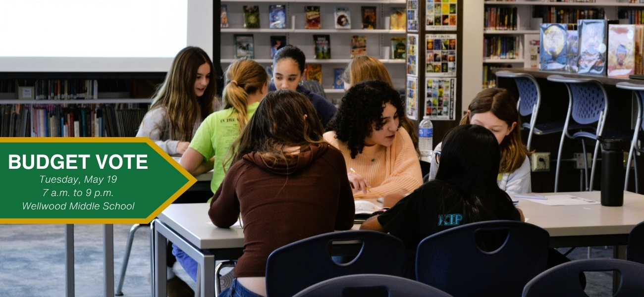 group of students at a table talking; text on slie says "budget Vote, Tuesday, may 19, 7 a.m. to 9 p.m., Wellwood Middle School"