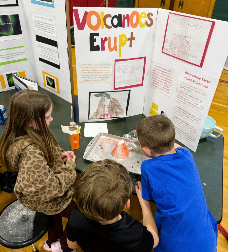Three students look at science fair project board on volcanoes.