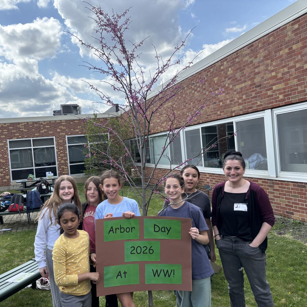 Students standing in font f blooming tree holding a sign saying "Arbor Day 2026 at Wellwood"