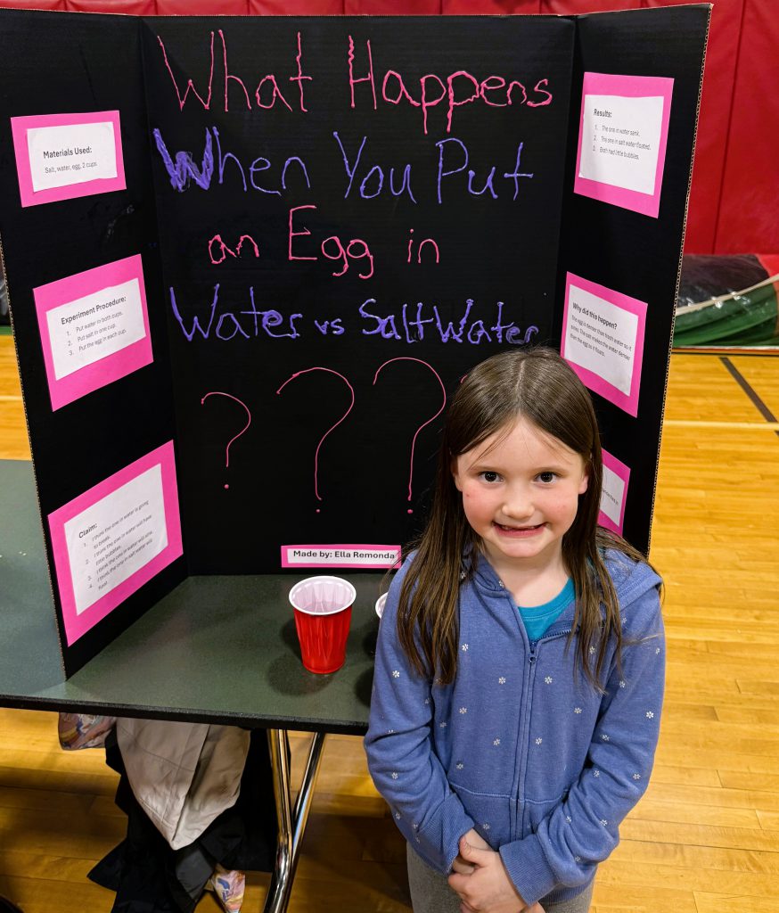Student stands in front of tri-fold display about what happens when you put an egg in water vs. salt water.