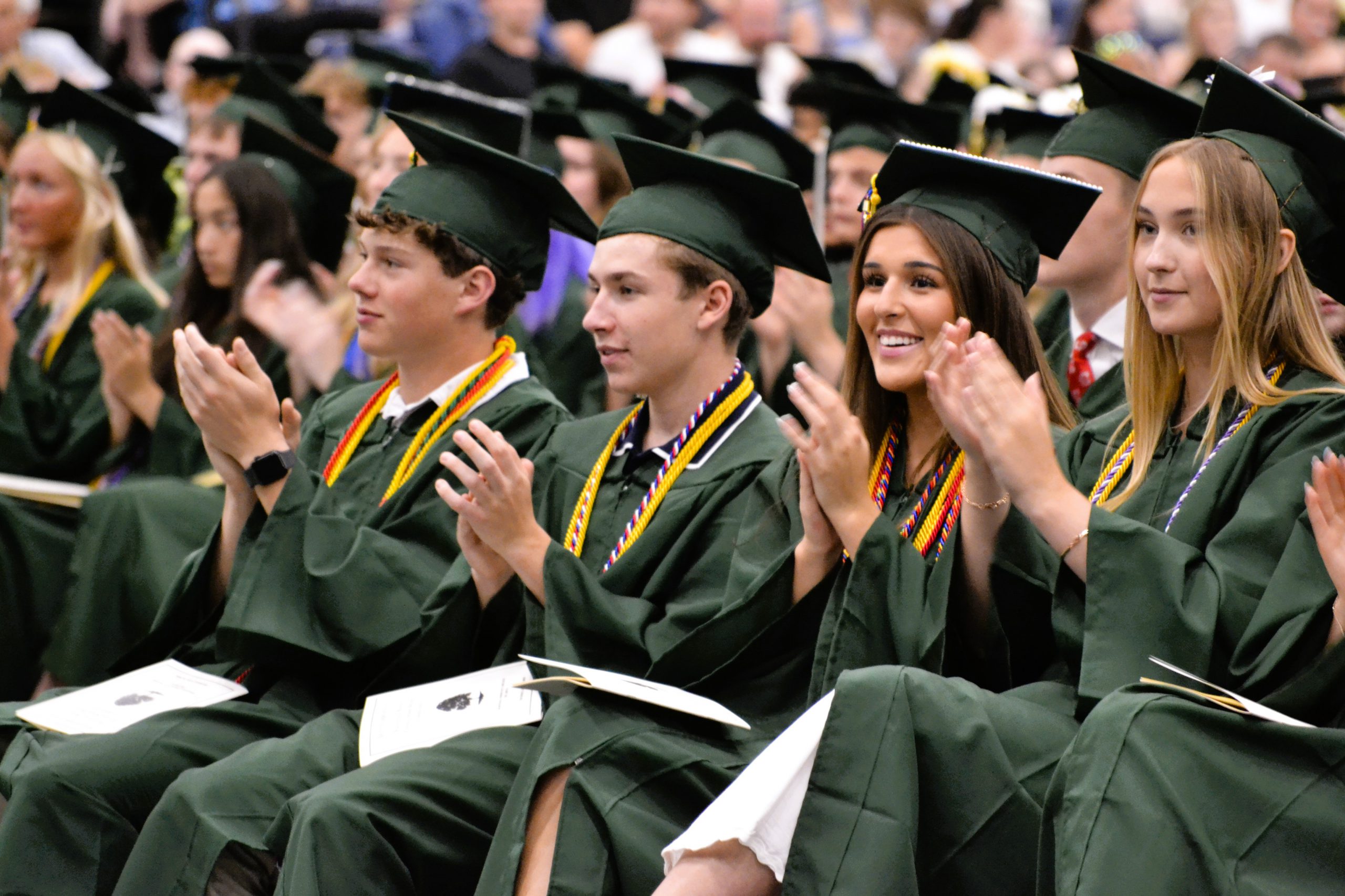 Students clap during graduation ceremony while wearing green caps and gowns.