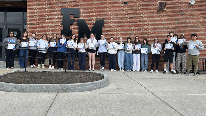 Students standing in a row, side by side, outside in front of a building, holding paper certificates in front of them