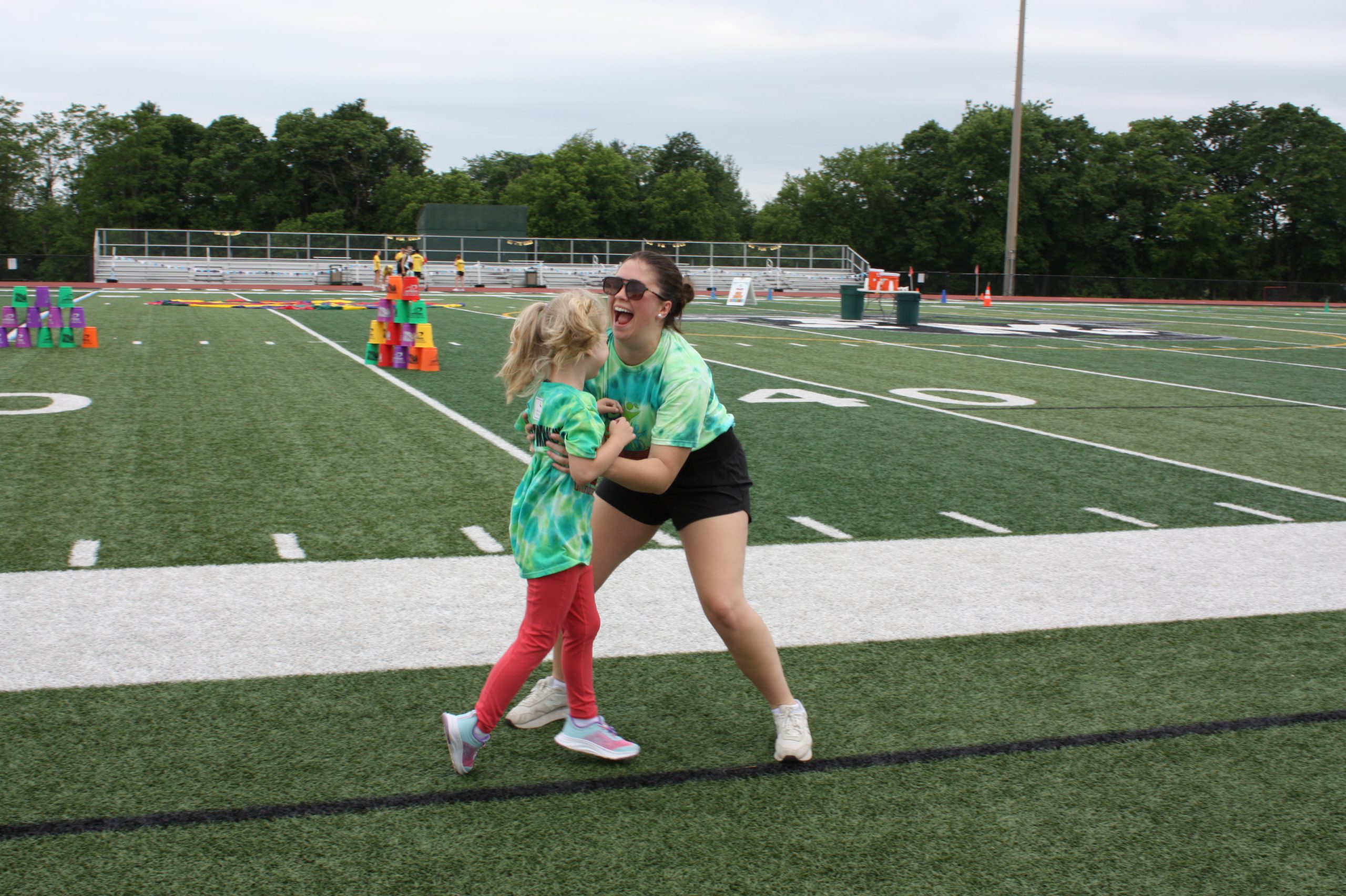 woman wearing shorts, sunglasses  and t-shirt catches a running student under the students arms, the student is facing the teacher and the teacher is laughing with her mouth opening. They are standing on the side of the turf field. 