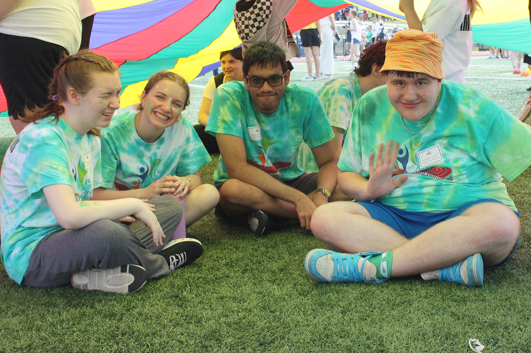 Four students seated under a rainbow colored parachute being held up by others- the photo really focuses on the four students who are seated on the ground, side by side, looking at the camera.