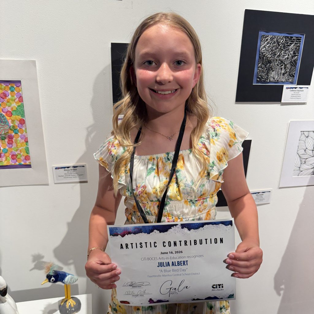 Girl standing in front of a white wall with artwork hanging on it; she's holding a certificate across the front of her body