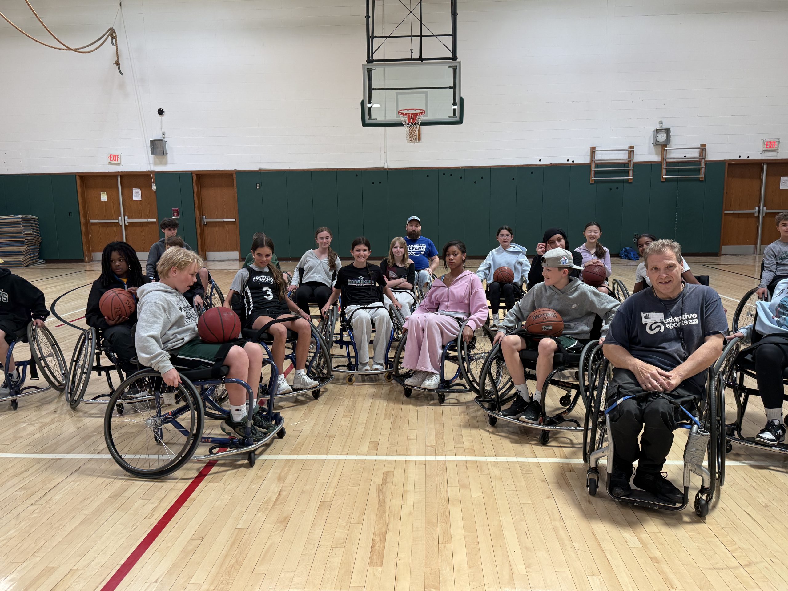 Gym class students sit in wheelchairs for adaptive sports lesson.